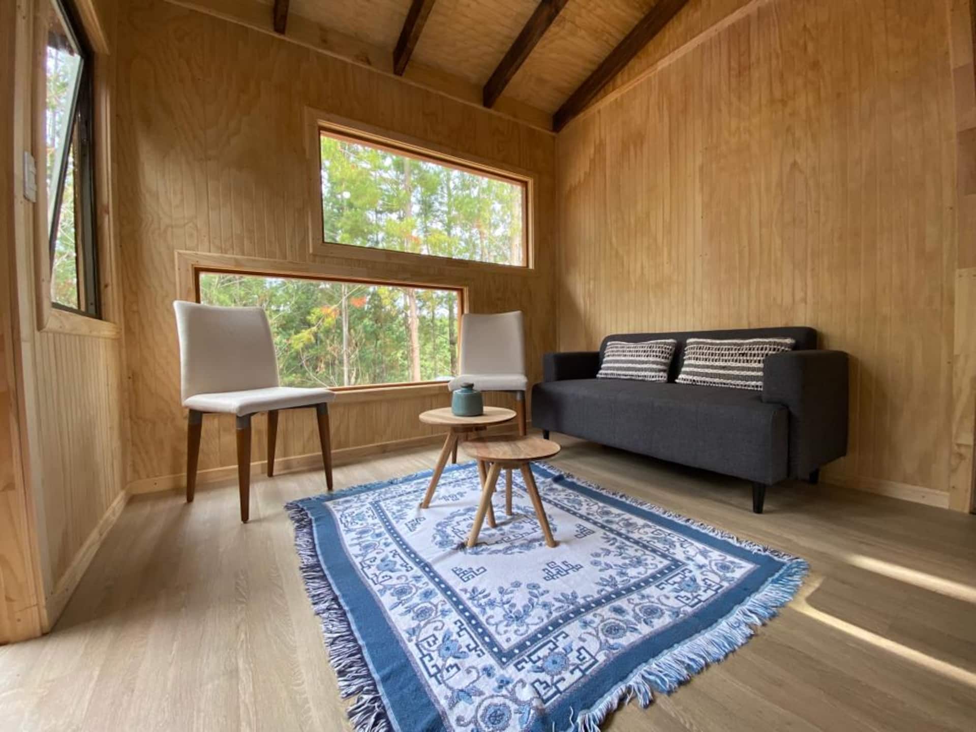 Living de la cabaña con sofá, dos sillas, mesa baja y un ventanal que mira al bosque